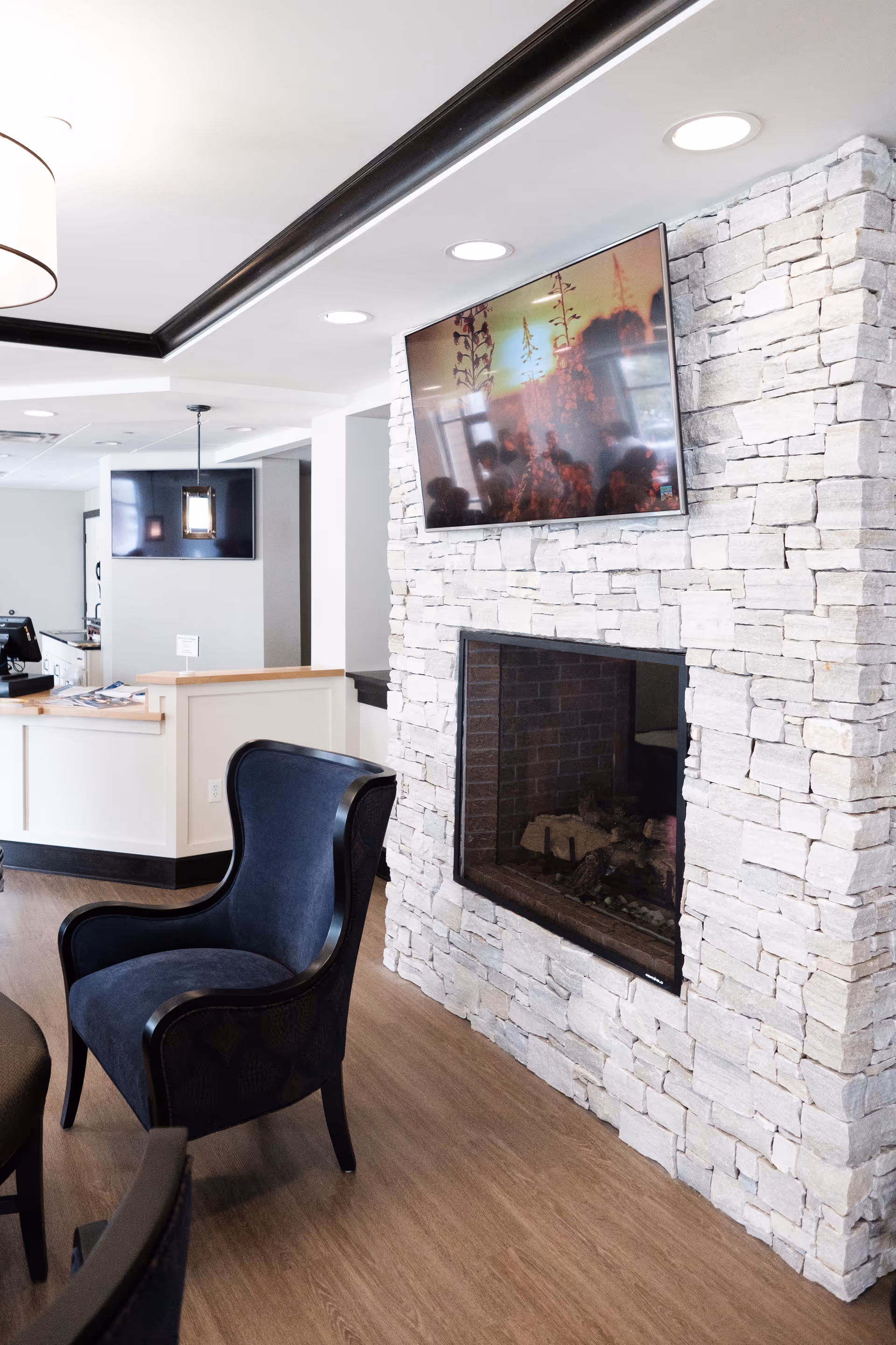 Modern lounge area with a stone fireplace, wall-mounted TV, and a navy upholstered armchair near a reception desk.