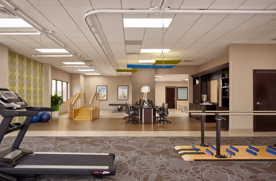 A spacious rehabilitation room at Alta Rehab at Fairmont featuring a treadmill on the left, parallel bars for walking exercises on the right, a set of stairs with handrails in the background, and a central area with desks and chairs equipped with computers. The room has a patterned carpet, wood flooring, and neutral-colored walls with framed artwork and modern ceiling lights.