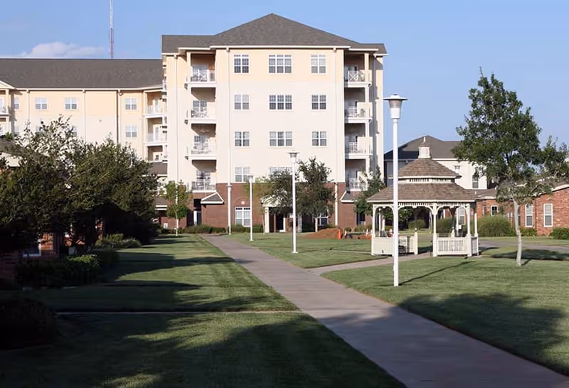A senior living facility with a multi-story beige building in the background, a paved walkway lined with lamp posts, green lawns, trees, and a white gazebo on the right side under a clear blue sky.