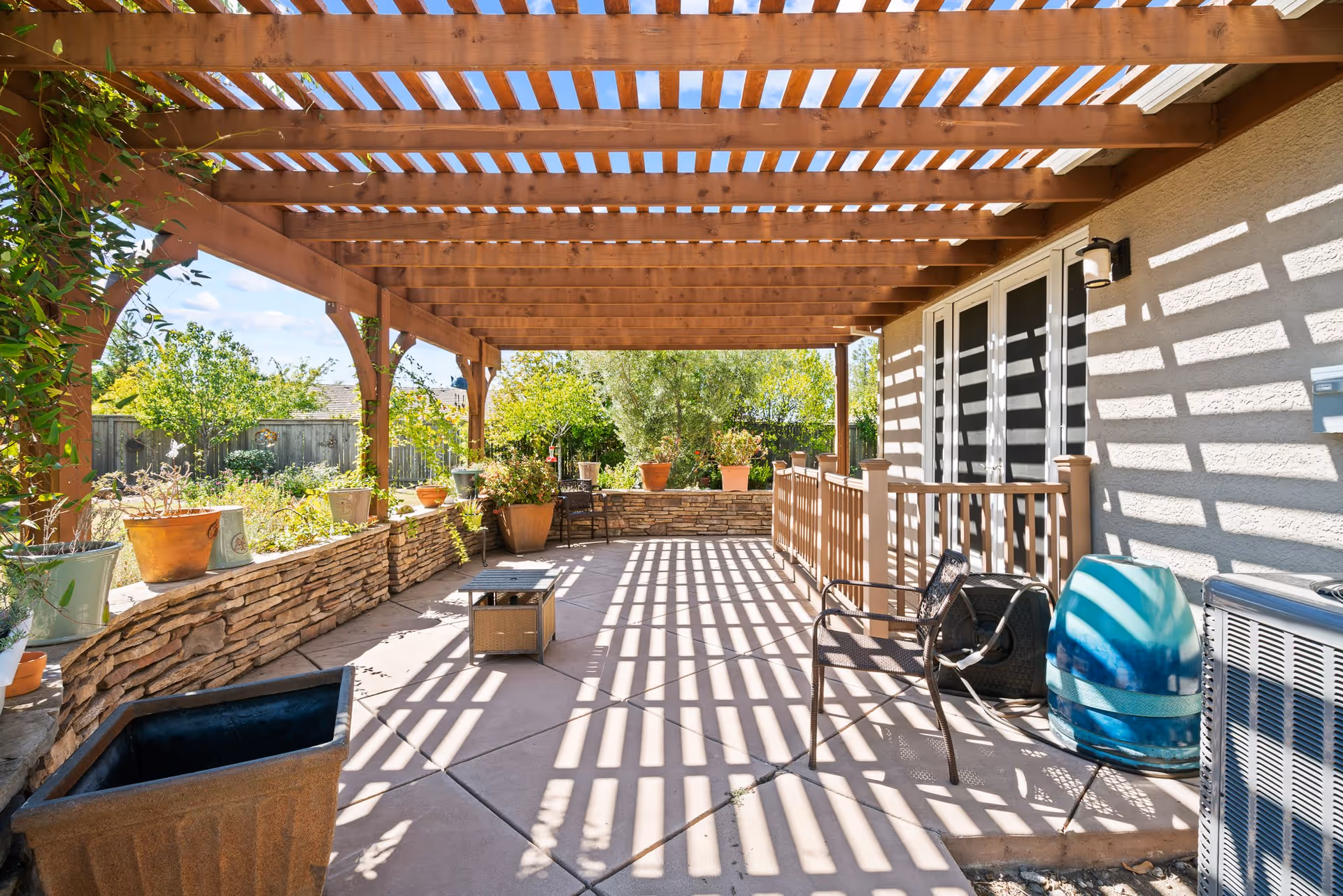 A shaded outdoor patio area with a wooden pergola casting striped shadows on the concrete floor. The patio is decorated with various potted plants along a stone wall, a small table, and a couple of chairs. The area is adjacent to a building with a light-colored exterior wall and glass doors.
