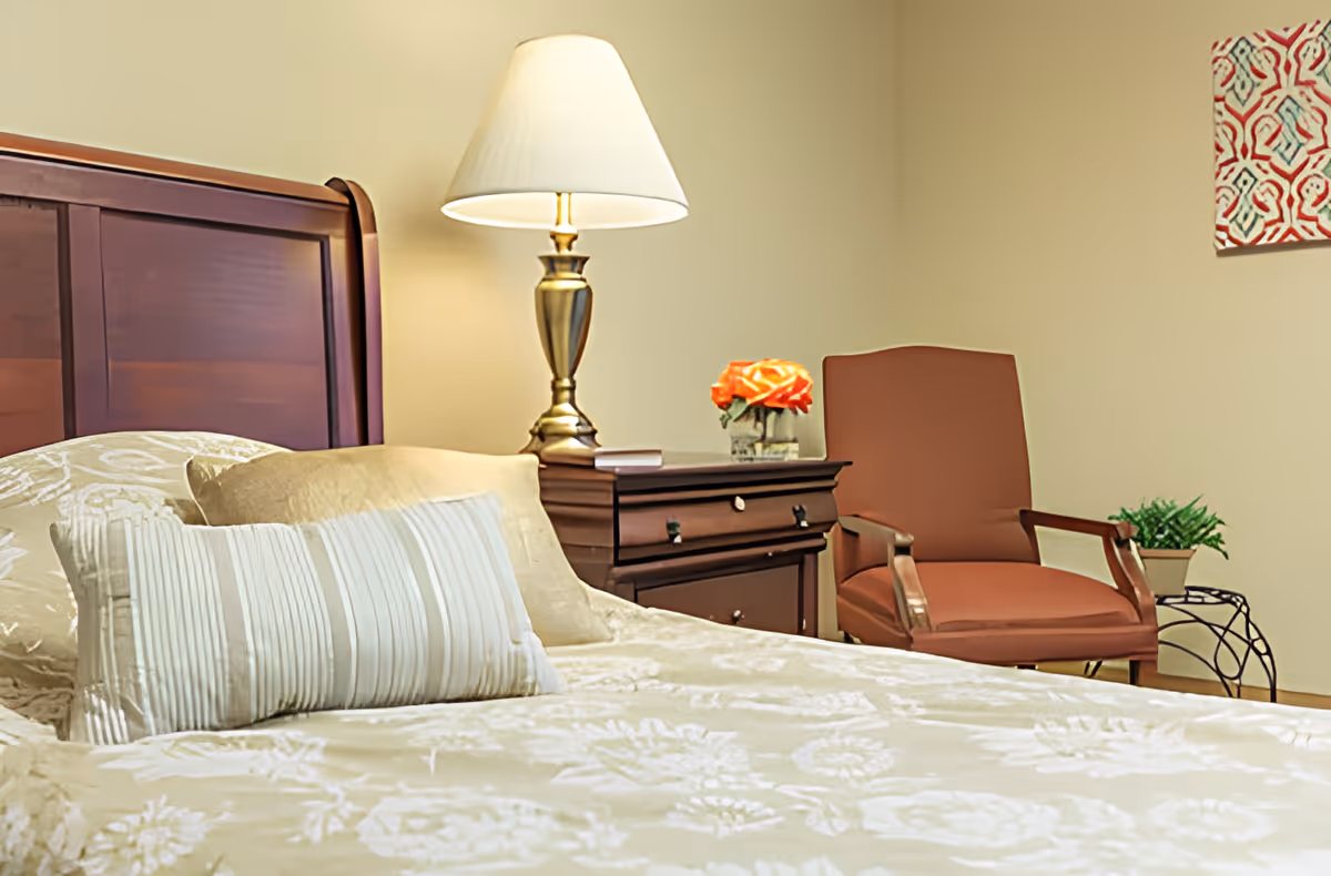 A cozy bedroom featuring a wooden headboard, a bed with cream-colored floral bedding and striped pillows, a wooden nightstand with a brass lamp and a small vase of orange flowers, a brown upholstered armchair, and a small side table with a green plant. A colorful patterned artwork is partially visible on the wall.