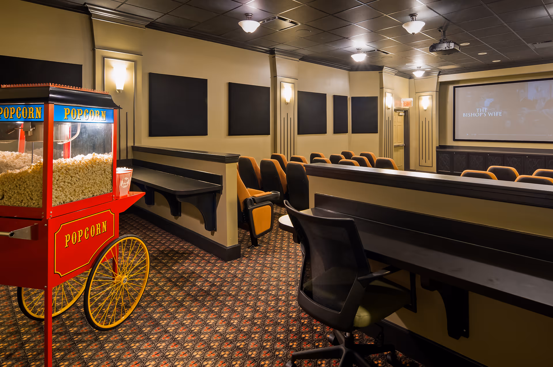 Interior of a small movie theater room with rows of cushioned seats facing a large screen displaying the title 'The Bishop's Wife'. A red popcorn cart filled with popcorn is positioned on the left side of the room. The room has patterned carpet, beige walls with black acoustic panels, and ceiling lights.