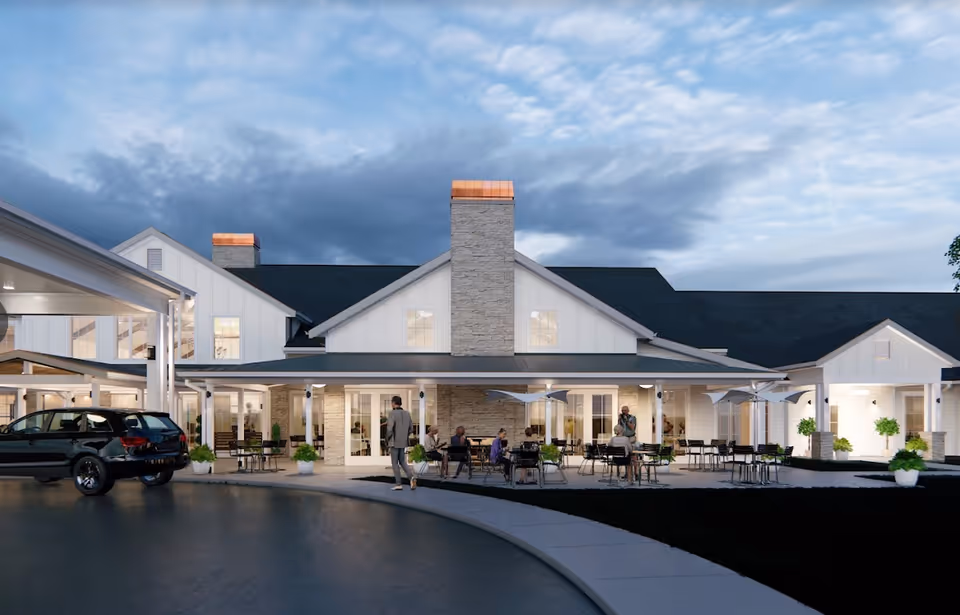 Exterior view of Cedarhurst Senior Living of Topeka at dusk, showing a modern building with white siding, large windows, and a stone chimney. There are outdoor seating areas with tables, chairs, and umbrellas where several people are sitting and standing. A black SUV is parked near the entrance under a covered driveway.