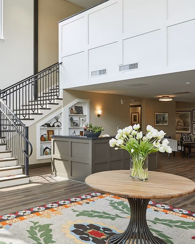Interior view of a senior living facility lobby area with a round wooden table holding a vase of white tulips in the foreground. Behind the table is a gray reception desk with a potted plant on top. To the left, there is a staircase with black metal railings and built-in shelves underneath displaying decorative items. The floor has a colorful patterned rug, and the walls are painted in neutral tones with framed artwork and wall sconces.