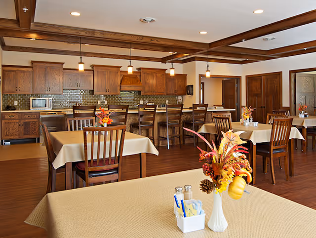 A dining area with several tables covered in beige tablecloths, each decorated with a fall-themed floral centerpiece. The room features wooden chairs and a kitchen area in the background with wooden cabinets, a microwave, and a tiled backsplash. Pendant lights hang from the ceiling, illuminating the space.