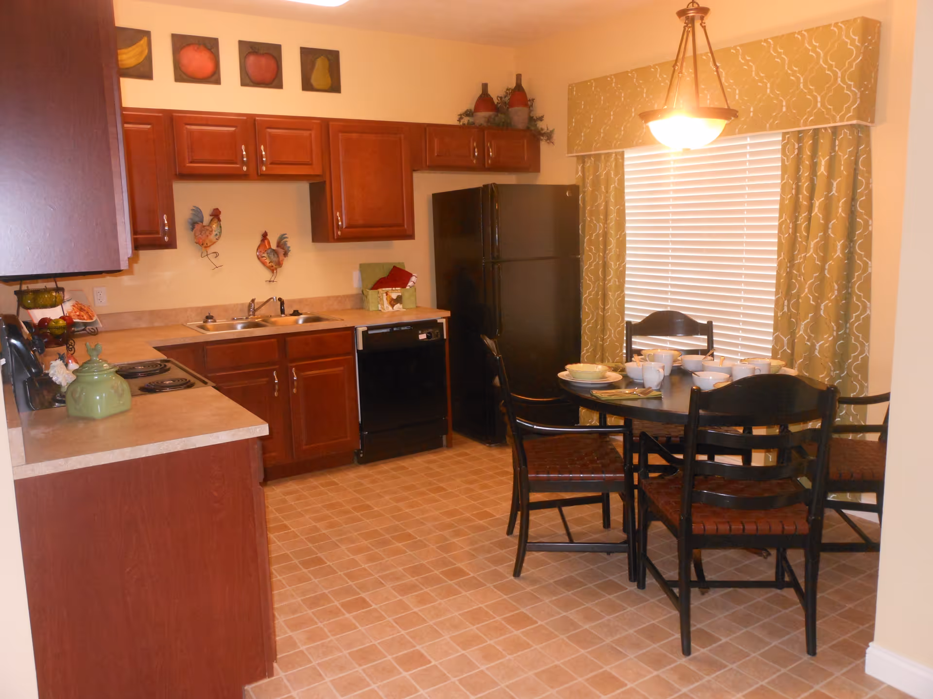 A kitchen and dining area with wooden cabinets, a black refrigerator, a black dishwasher, and a double sink. There is a round dining table with four chairs, set with bowls, cups, and plates. The window behind the table has blinds and patterned curtains. The walls are decorated with fruit paintings and rooster wall hangings.