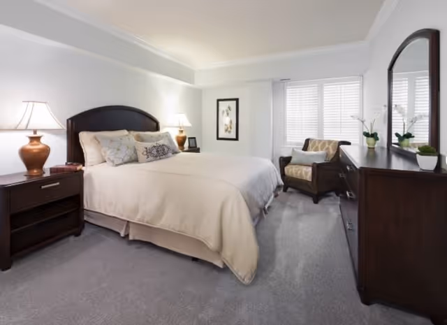 A neatly made bed with beige bedding and multiple pillows in a bright bedroom. The room features two bedside tables with lamps, a framed picture on the wall, a cushioned armchair near a window with blinds, and a dark wooden dresser with a mirror and potted plants on top.