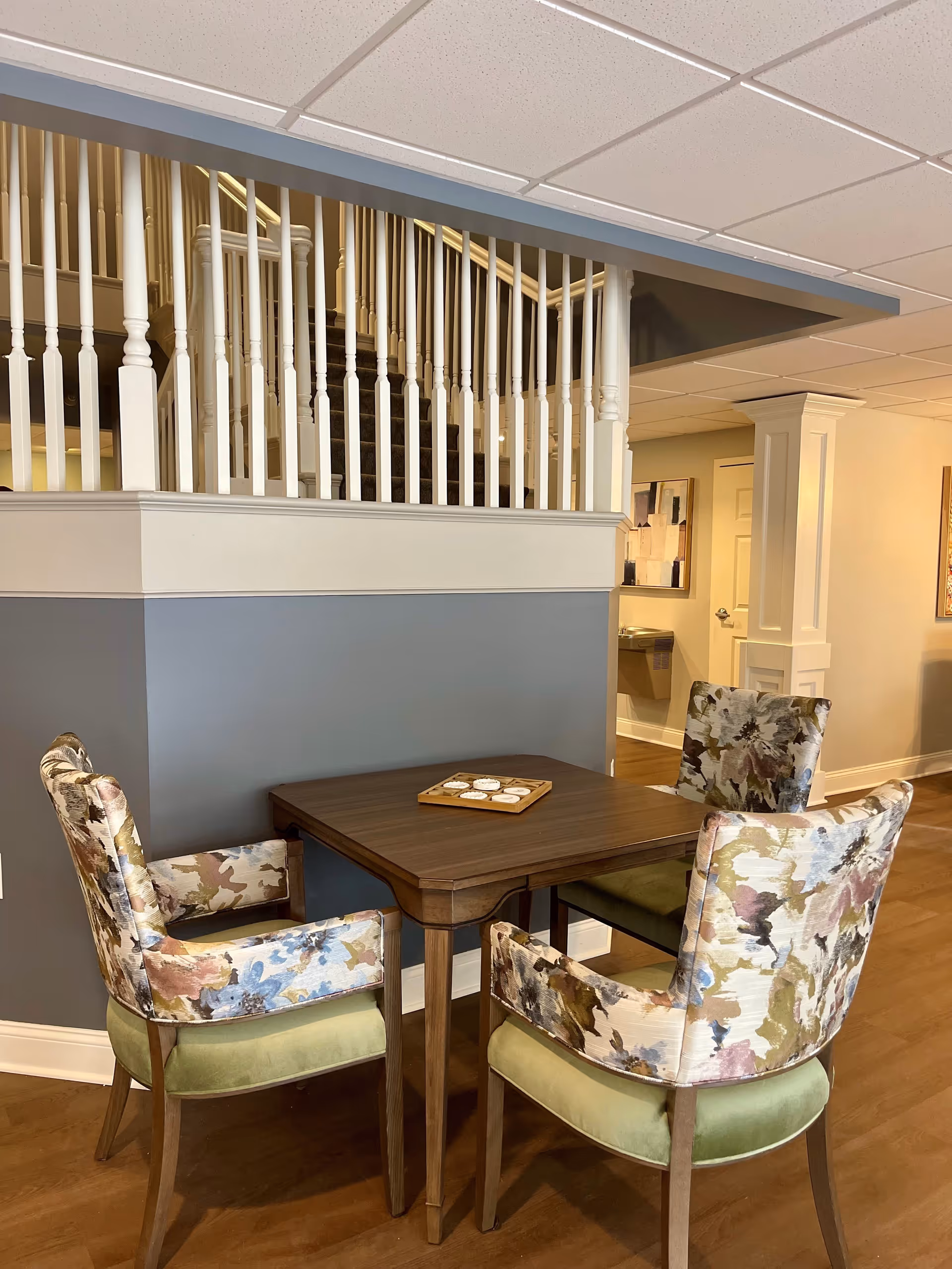 A small wooden table with a board game on top, surrounded by four upholstered chairs with floral patterned backs and green seats, set in a room with wooden flooring and a white railing above a half wall painted gray. In the background, there is a hallway with a water fountain and framed artwork on the walls.