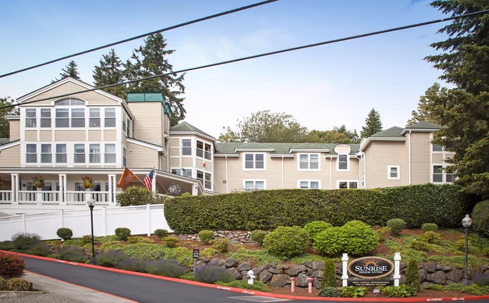Exterior view of a multi-story senior living facility building with beige siding and multiple windows. The building is surrounded by well-maintained landscaping including bushes, trees, and a stone retaining wall. There is a paved driveway with red curbs and a sign in front that reads 'Sunrise Senior Living'. Flags are displayed near the entrance, and the sky is clear with some light clouds.