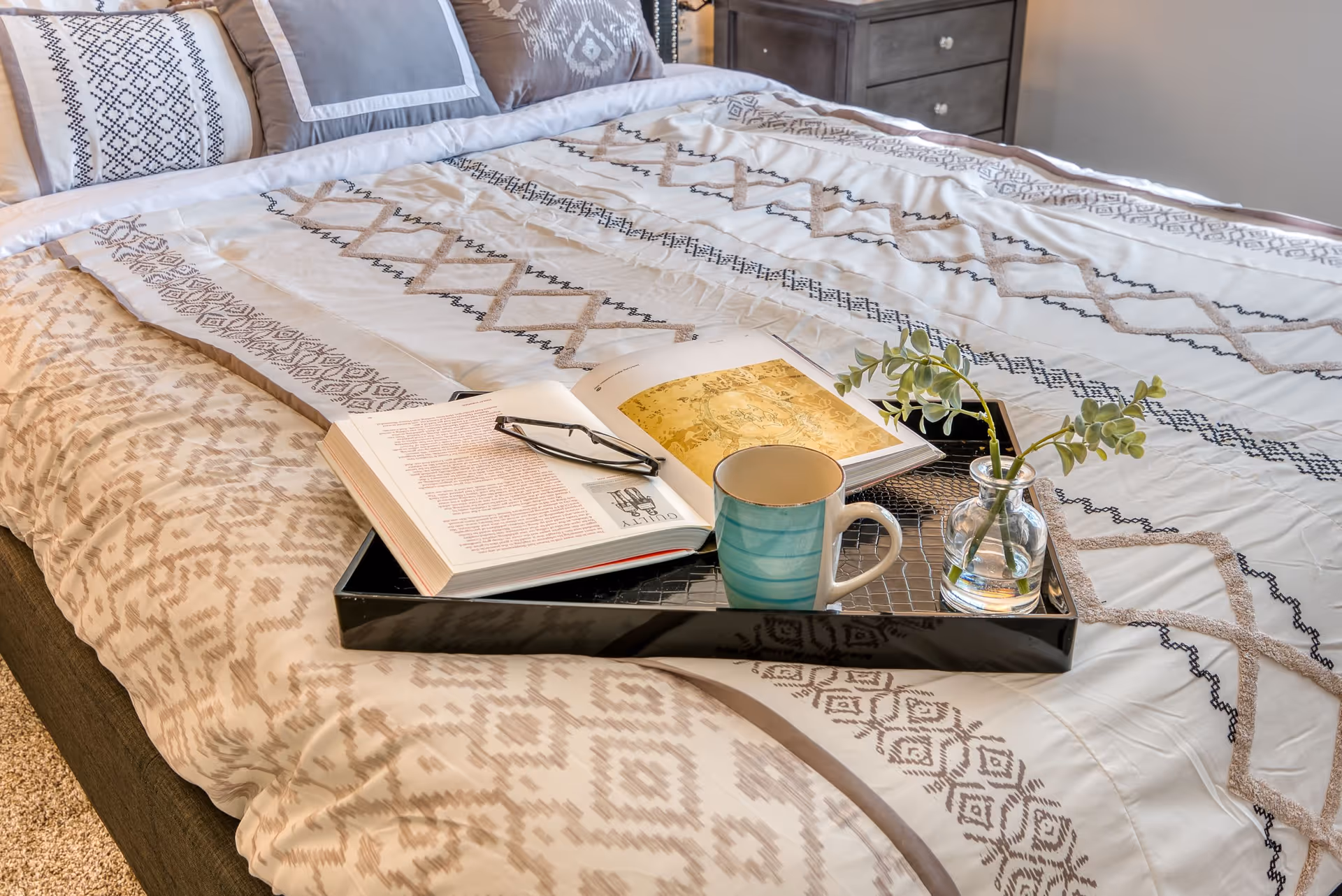 A neatly made bed with patterned bedding in neutral tones. On the bed is a black tray holding an open book, a pair of reading glasses, a blue-striped coffee mug, and a small glass vase with green leafy stems. A dark wooden nightstand with drawers is visible in the background.