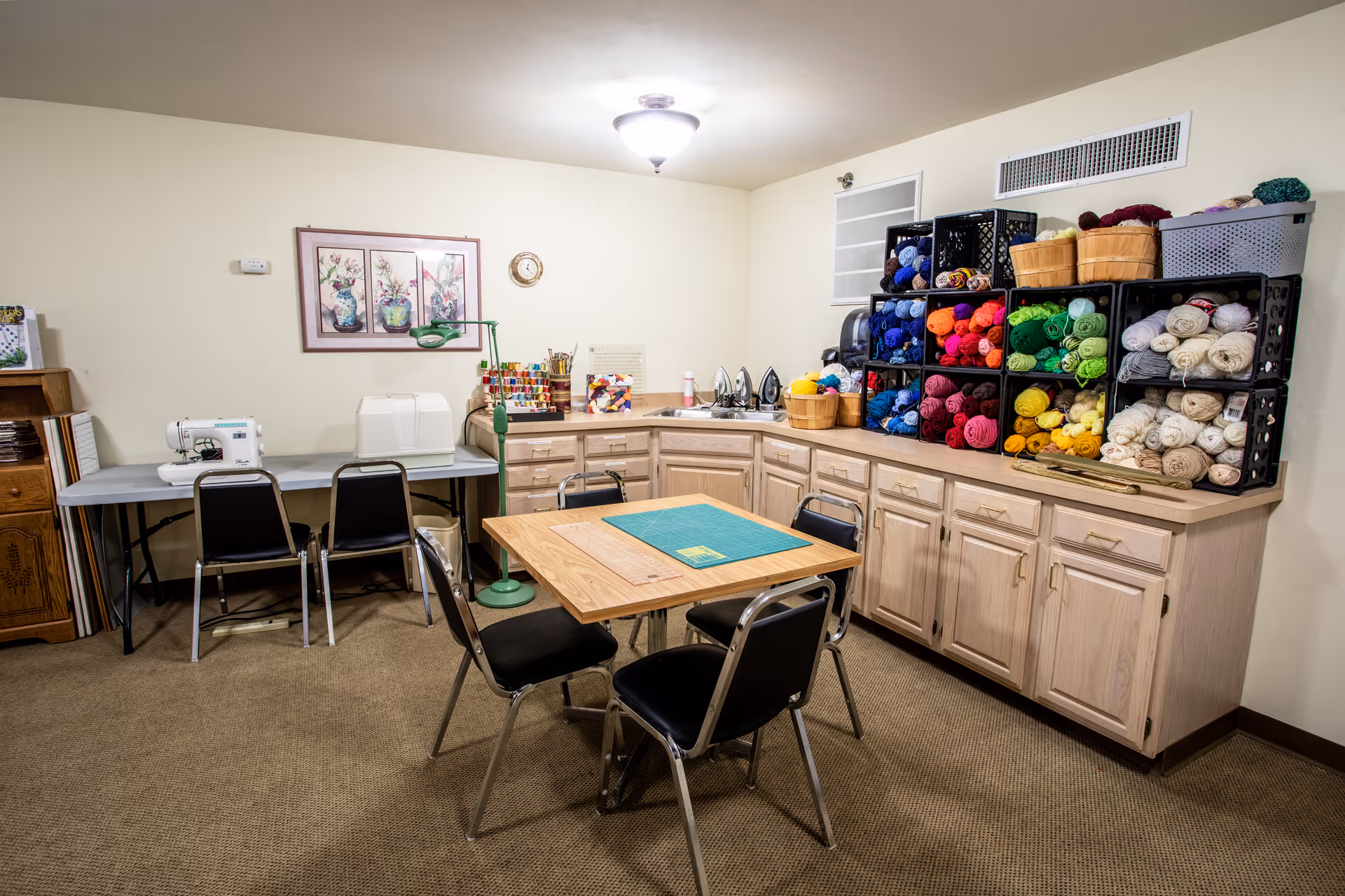 A craft room with a central table and chairs, sewing machines on a side table, and shelves filled with colorful yarn and supplies.