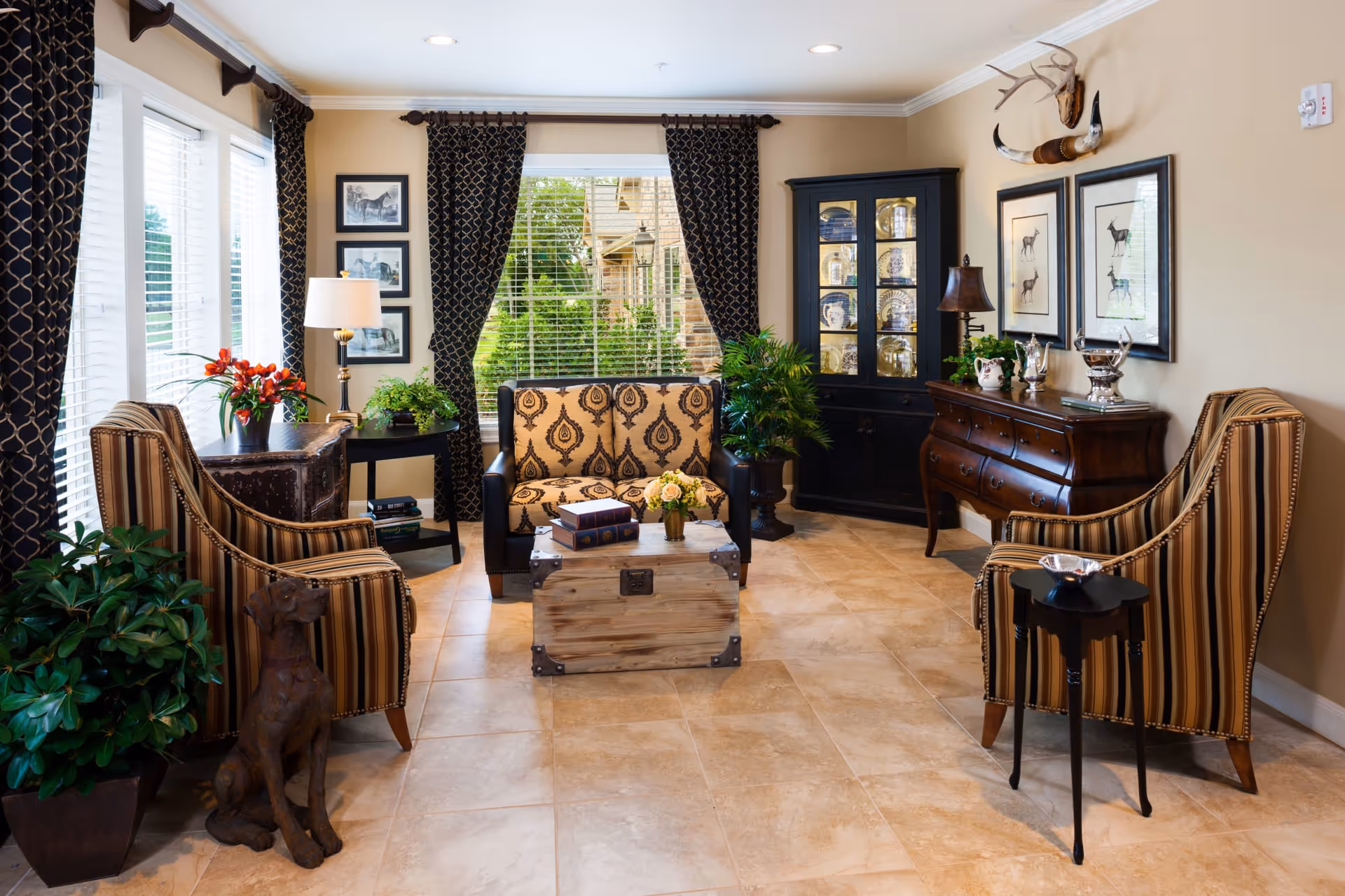 A cozy living room with beige tiled floors, two striped armchairs, a patterned loveseat, a wooden chest used as a coffee table, and various plants. The room features large windows with dark patterned curtains, framed animal artwork on the walls, a black display cabinet with glass doors, and a wooden sideboard with decorative items. A dog statue is placed near one of the armchairs.
