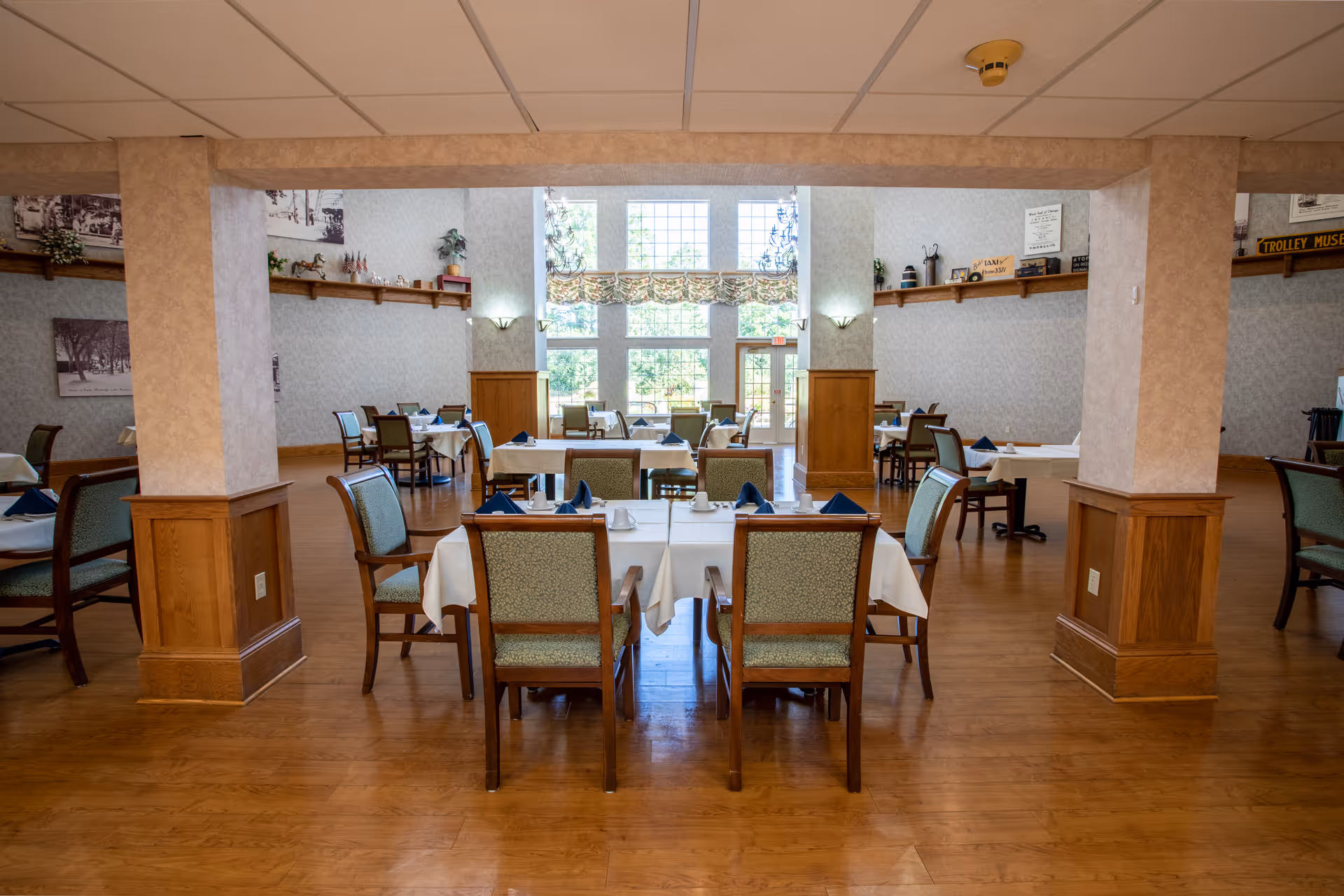 A spacious dining room in a senior living facility with wooden floors, multiple tables covered with white tablecloths, each set with cups and blue napkins. The room features large windows letting in natural light, beige walls with wooden paneling on the lower half, and decorative shelves with plants and ornaments.