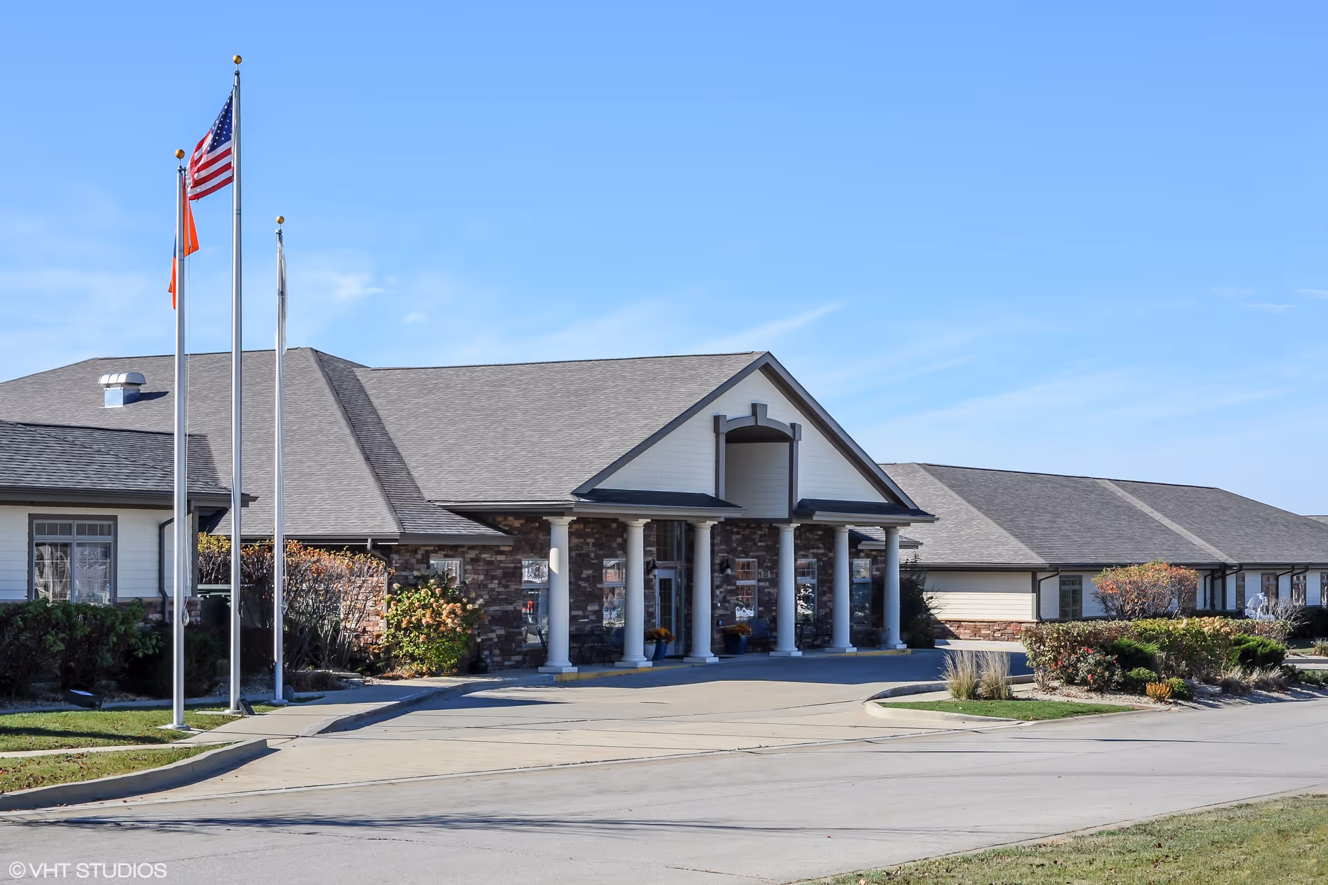Exterior view of Bridle Brook Assisted Living & Memory Care Community building with a driveway, three flagpoles with flags, and landscaped bushes under a clear blue sky.