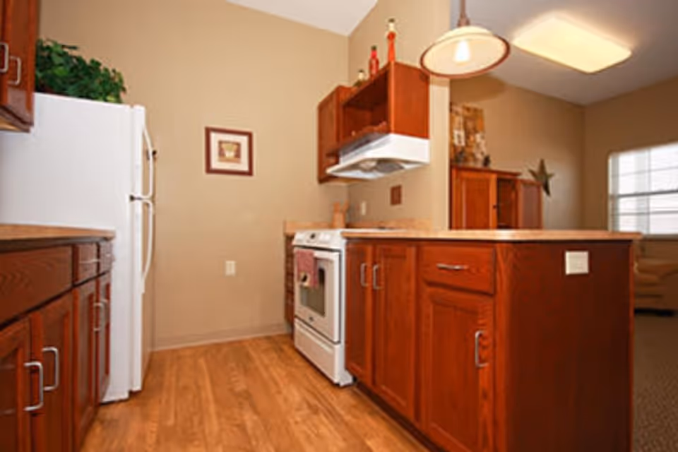 A kitchen with wooden cabinets and drawers, a white refrigerator, a white stove with an overhead range hood, and a wooden floor. There is a pendant light hanging above the counter and a window in the adjacent room letting in natural light.