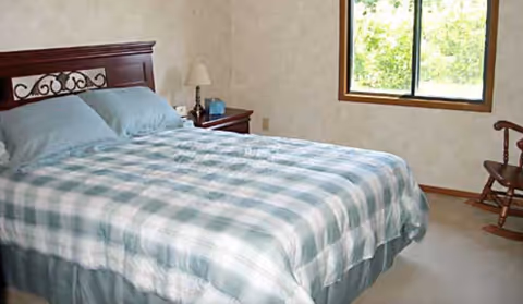 A bedroom with a neatly made bed featuring a blue and white checkered comforter and two blue pillows. There is a wooden headboard, a nightstand with a lamp and a clock, a window showing greenery outside, and a wooden rocking chair in the corner.