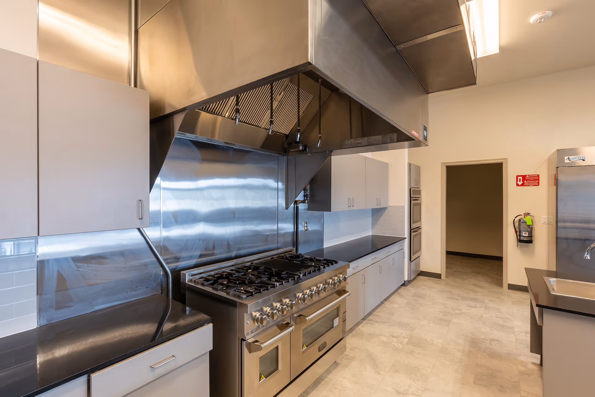 A commercial kitchen featuring a large stainless steel gas stove with multiple burners and ovens, a stainless steel ventilation hood above, light-colored cabinets with silver handles, a stainless steel backsplash, a double wall oven, a fire extinguisher mounted on the wall, and a stainless steel refrigerator. The floor is tiled with light-colored tiles and the walls are painted beige.