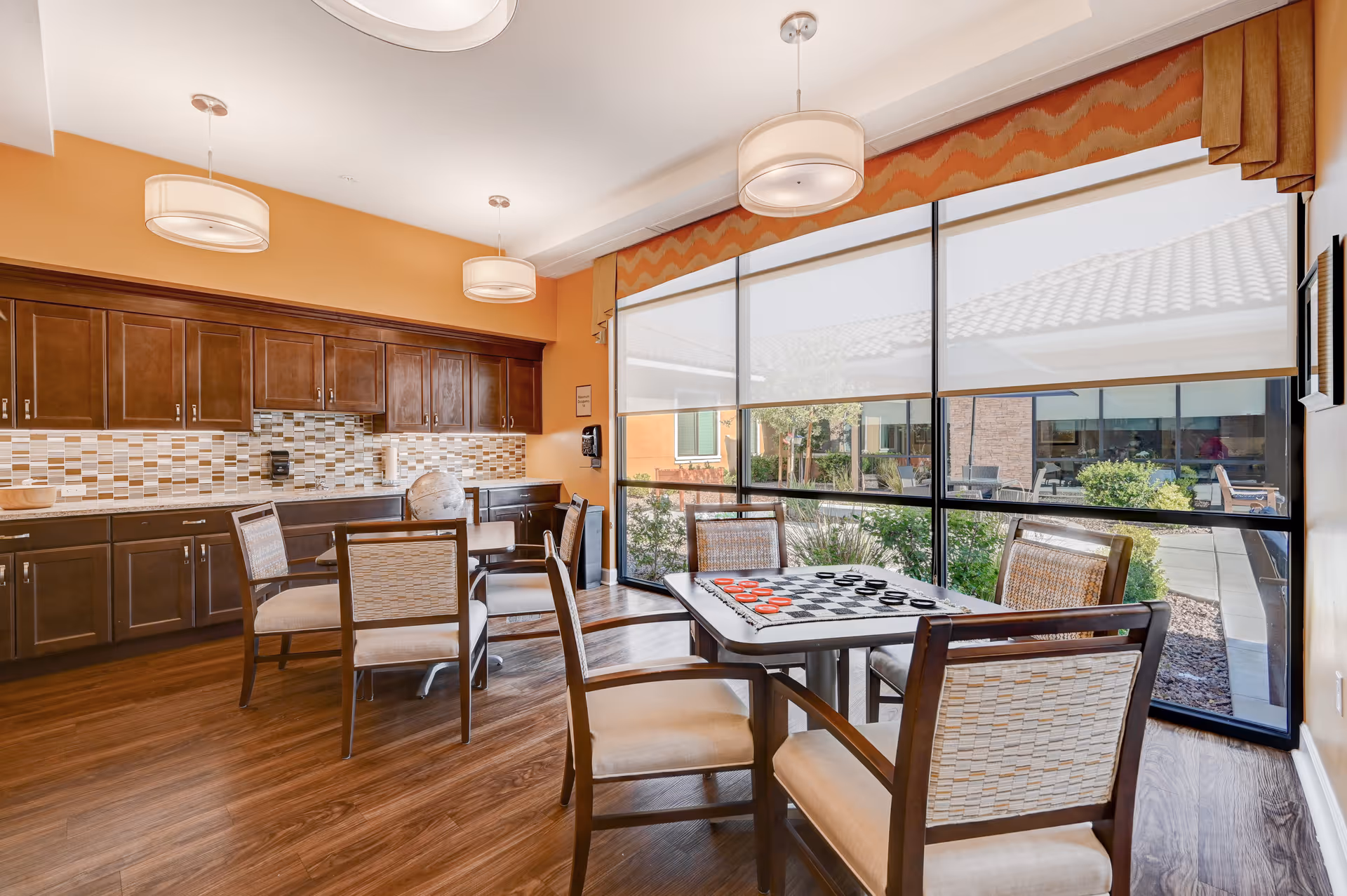 Sunlit dining and activity room with a kitchenette, tables and chairs, and a checkerboard by large windows overlooking a courtyard.