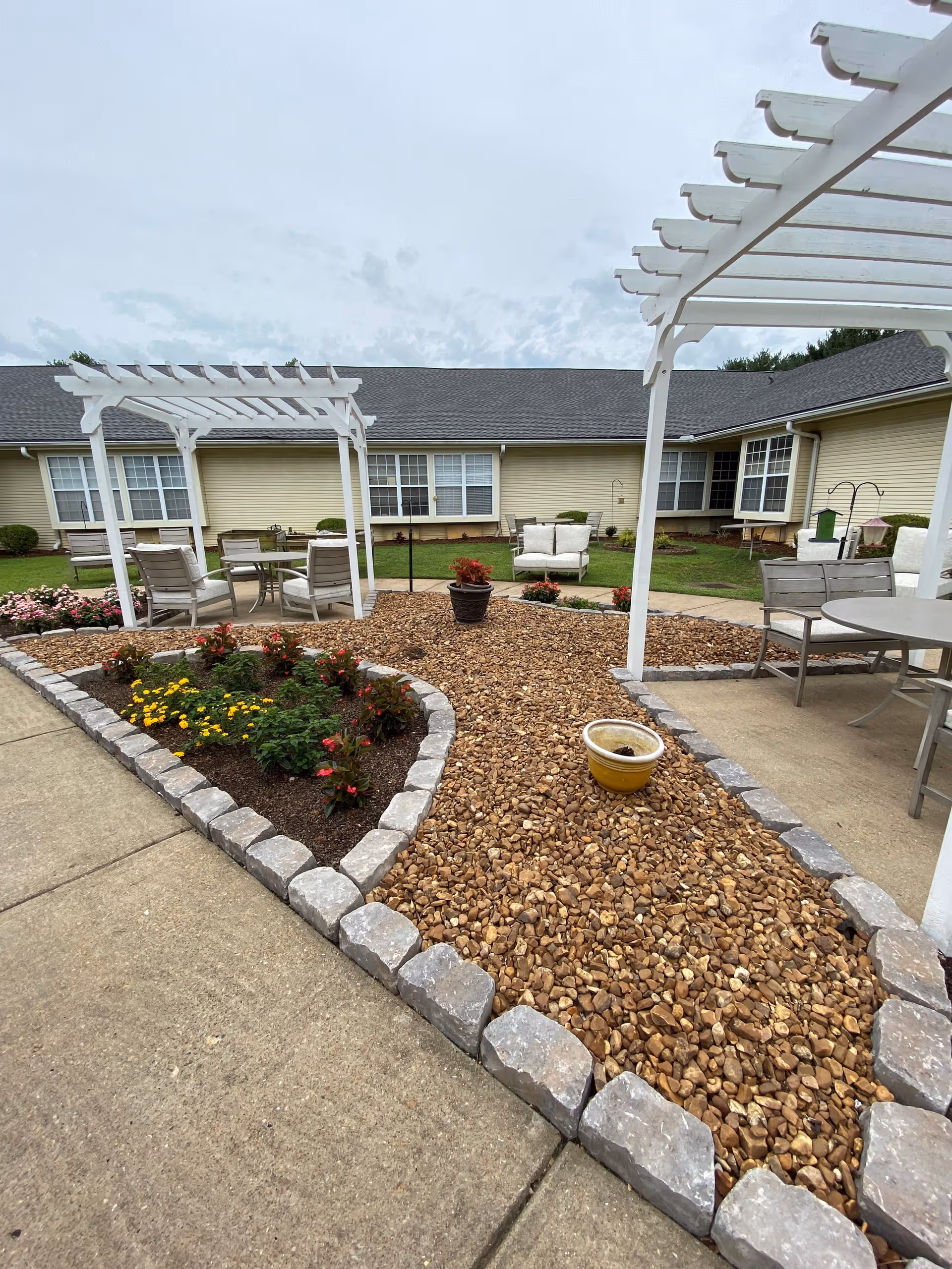 Outdoor patio area at American House Murfreesboro featuring two white pergolas, seating areas with chairs and tables, a gravel pathway bordered by stone, flower beds with blooming plants, and a beige building with multiple windows in the background under a cloudy sky.
