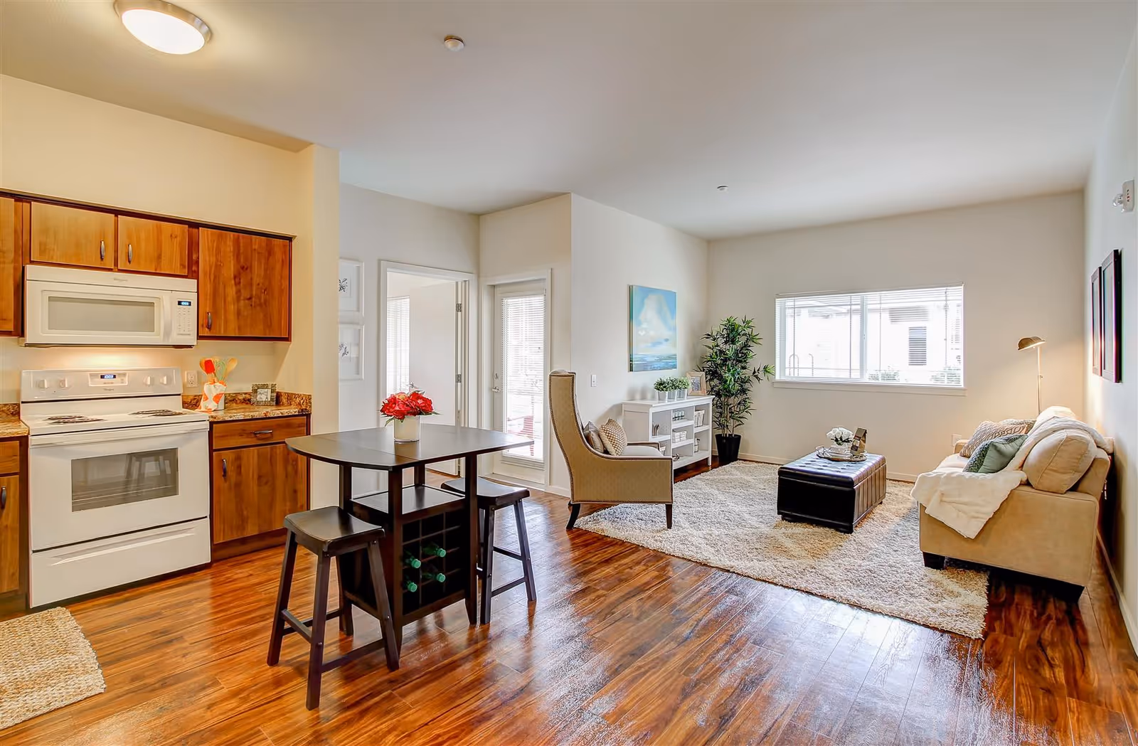A bright and cozy open-plan living space featuring a kitchen area with wooden cabinets, a white stove and microwave, and a small dining table with two stools. The living area has a beige sofa, an armchair, a white entertainment unit, a large window, a floor lamp, a potted plant, and a soft area rug on wooden flooring.