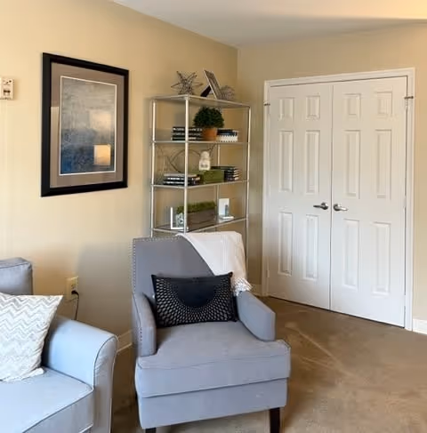 A cozy living room corner with a gray armchair featuring a black pillow and a white throw blanket. Next to it is a light gray sofa with a chevron-patterned pillow. Behind the armchair is a metal shelving unit with decorative items and books. A framed abstract artwork hangs on the beige wall, and there is a double white door with silver handles in the background.