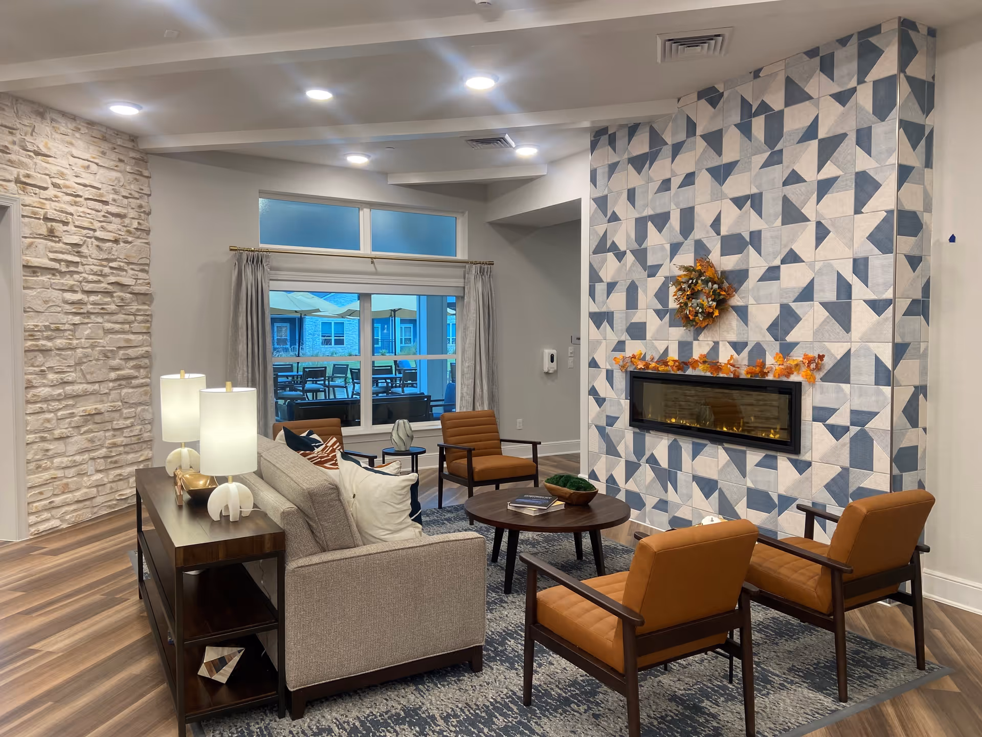 A modern living room with a gray sofa, two brown armchairs, and a round wooden coffee table on a blue patterned rug. The room features a decorative fireplace with a geometric blue and white tile surround, adorned with autumn leaves and a wreath. There are two table lamps on a wooden console behind the sofa, a stone accent wall on the left, and a large window with curtains showing an outdoor patio area with tables and umbrellas.