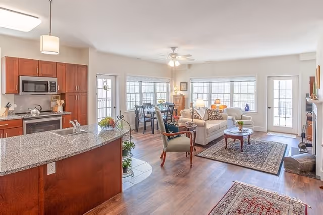 Bright and spacious open-concept living area in a senior living facility featuring a kitchen with granite countertops and wooden cabinets, a dining table with chairs near large windows, and a cozy living room with sofas, armchairs, a coffee table, and a fireplace.