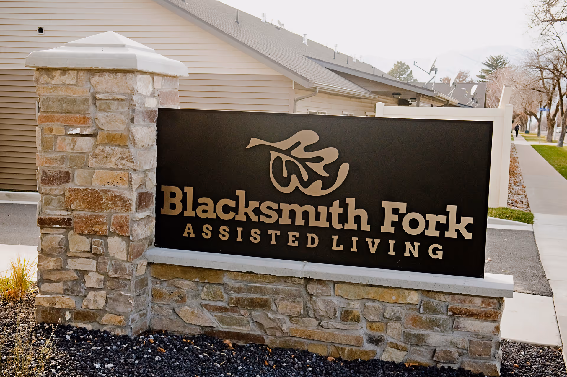 Stone and brick signpost with a black plaque displaying the name 'Blacksmith Fork Assisted Living' in beige letters, located outdoors near a sidewalk and residential buildings.