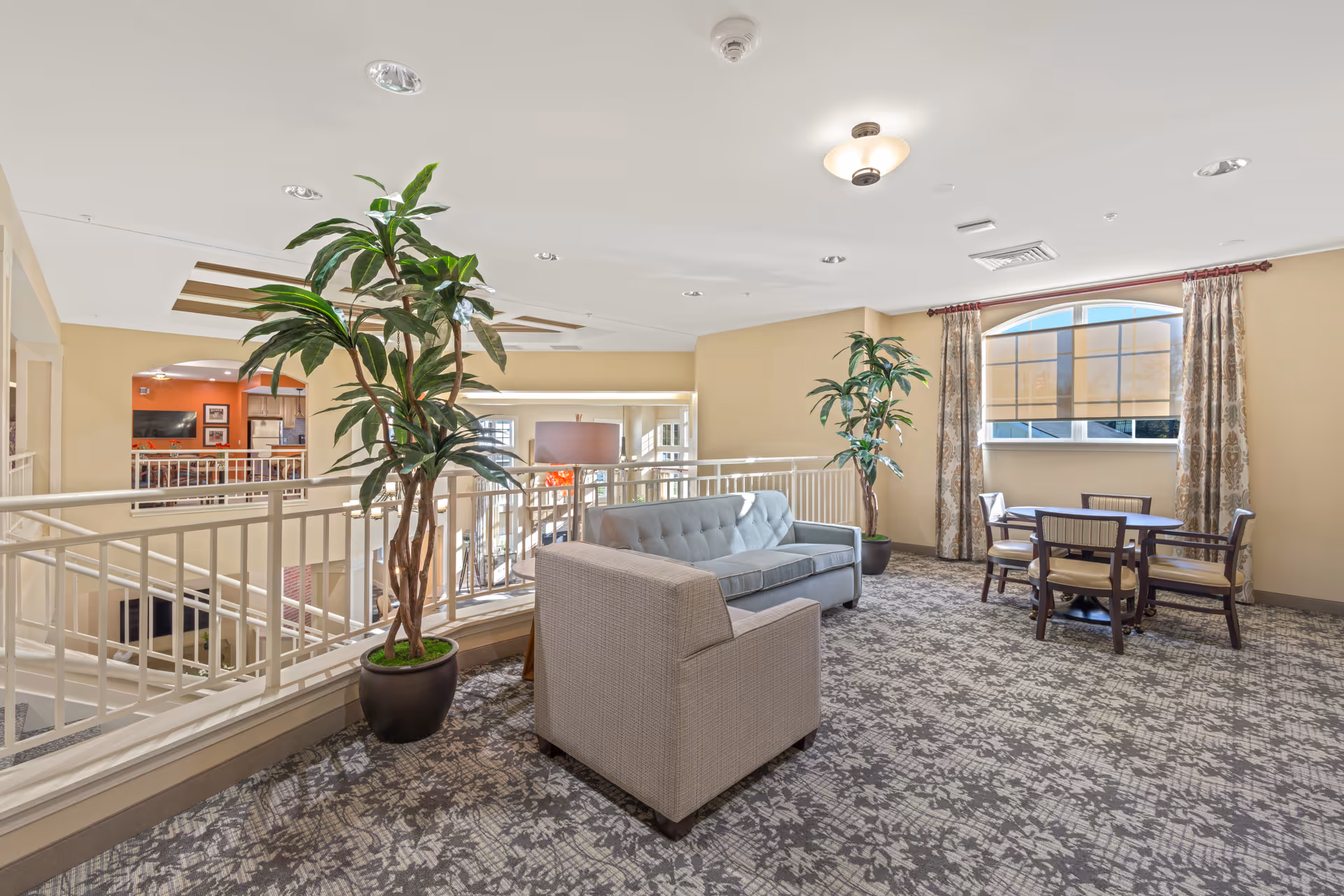 A bright and spacious interior common area with a light blue sofa, a beige armchair, two large potted plants, and a round wooden table with four chairs near a large window with patterned curtains. The area overlooks a lower level with railings and has a patterned carpet floor.