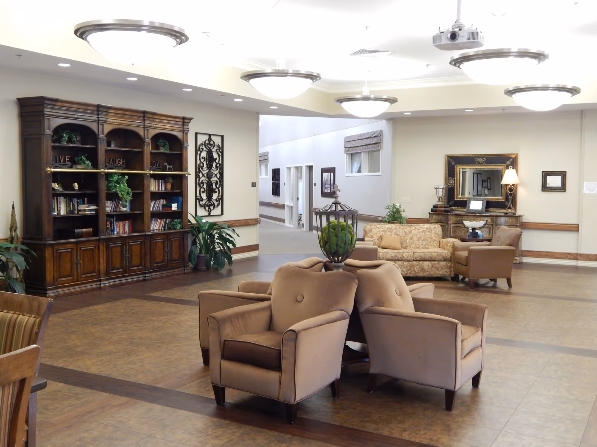 A spacious and well-lit common area in a senior living facility featuring comfortable armchairs arranged around a central decorative piece, a wooden bookshelf filled with books and plants, a floral-patterned sofa, an armchair, a wooden sideboard with a mirror and lamp, and decorative wall art. The floor is tiled and the ceiling has multiple round light fixtures.