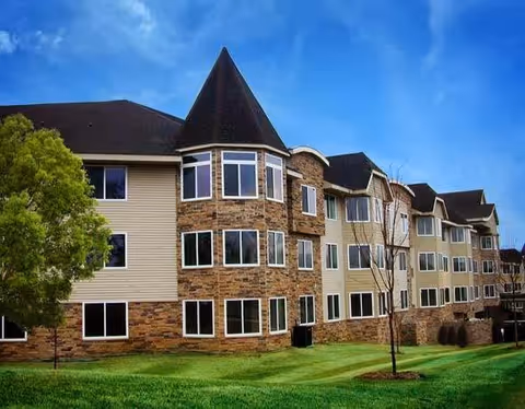 Exterior view of a multi-story senior living facility building with a combination of beige siding and stone facade, featuring multiple windows and a pointed turret-like architectural element under a blue sky.