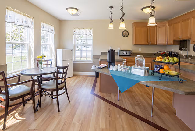 Sunny communal dining area and kitchenette with a round table and chairs, cabinets and windows, and a serving counter set with coffee, glasses, and fruit.