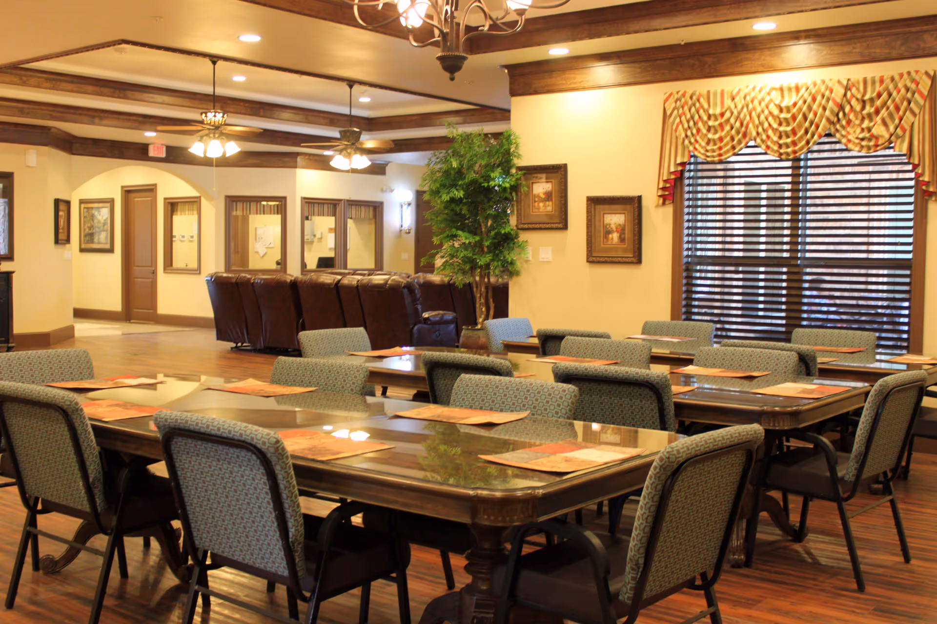 A dining room in Avalon Memory Care featuring several tables with placemats and cushioned chairs arranged neatly. The room has warm lighting, wooden floors, decorative window valances, framed artwork on the walls, and a potted plant near the back. In the background, there is a seating area with brown leather chairs and ceiling fans.