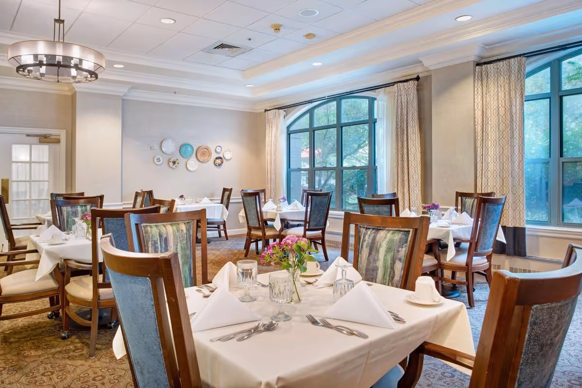 A bright and elegant dining room with several tables covered in white tablecloths, each set with folded napkins, glassware, and silverware. The room features large arched windows with patterned curtains allowing natural light to fill the space. Wooden chairs with cushioned seats surround the tables, and decorative plates are displayed on one wall. A modern chandelier hangs from the ceiling.