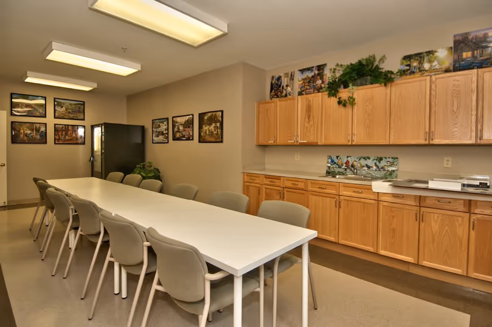 A long white table surrounded by beige chairs in a room with wooden cabinets along one wall. The cabinets have plants and pictures on top, and there are framed pictures hanging on the opposite wall. The room is well-lit with ceiling lights and has a refrigerator in the corner.