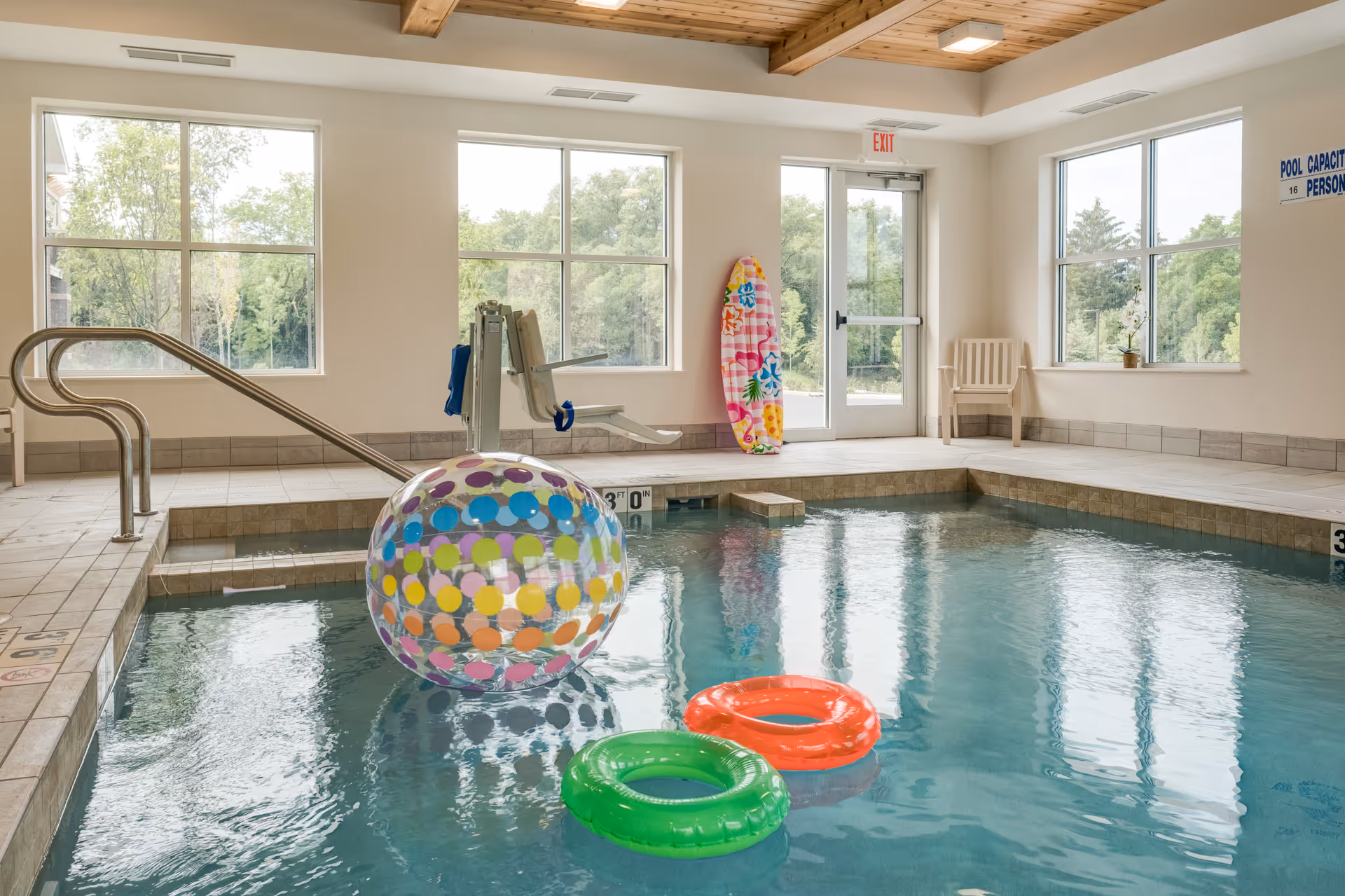 Indoor swimming pool area with large windows showing green trees outside. The pool has a colorful inflatable beach ball and two inflatable rings, one green and one orange, floating on the water. There is a pool lift chair for accessibility, a metal handrail, a colorful surfboard leaning against the wall, and a plastic chair near the exit door. The ceiling has wooden beams and the walls are light-colored.