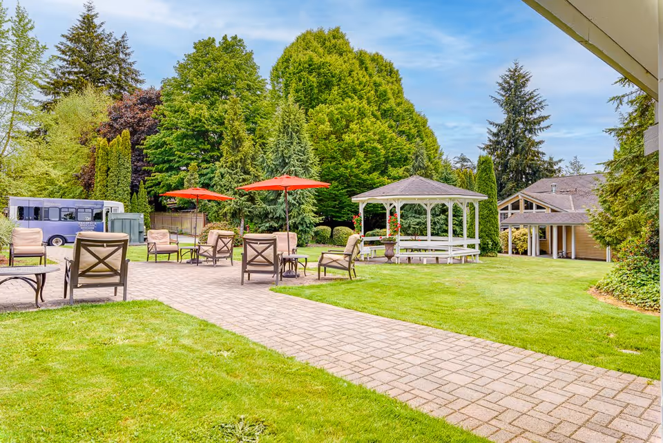 Outdoor patio area with several cushioned chairs and tables under red umbrellas, a white gazebo with flowers, lush green grass, and tall trees surrounding the space. A building and a shuttle bus are visible in the background under a partly cloudy blue sky.