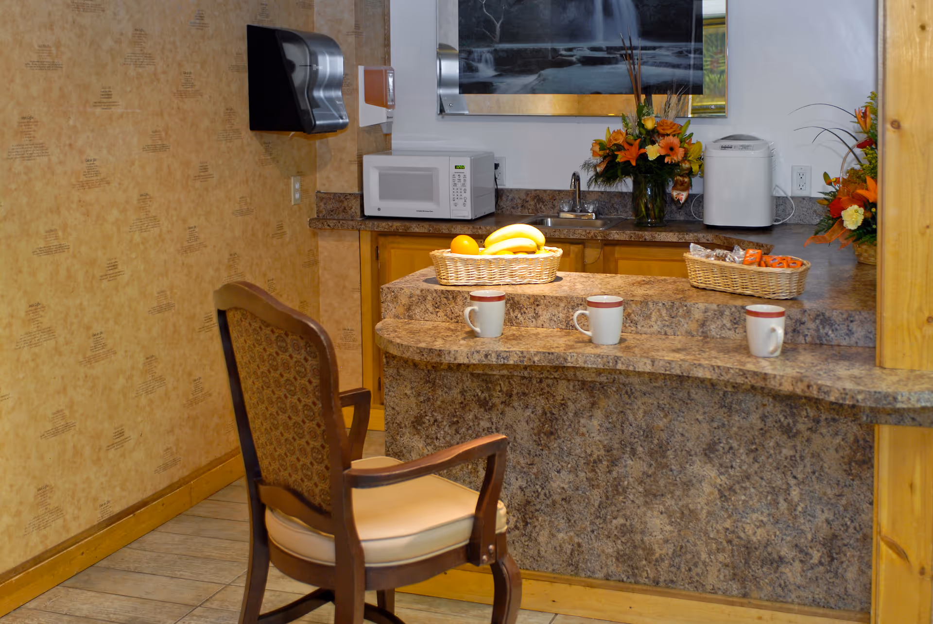 A small kitchen area with a granite countertop bar featuring three coffee mugs, a basket of bananas and oranges, and another basket with snacks. There is a wooden chair with a cushioned seat in front of the bar. On the back counter, there is a microwave, a small sink, a bread maker, and two flower arrangements. The walls have a beige patterned wallpaper.