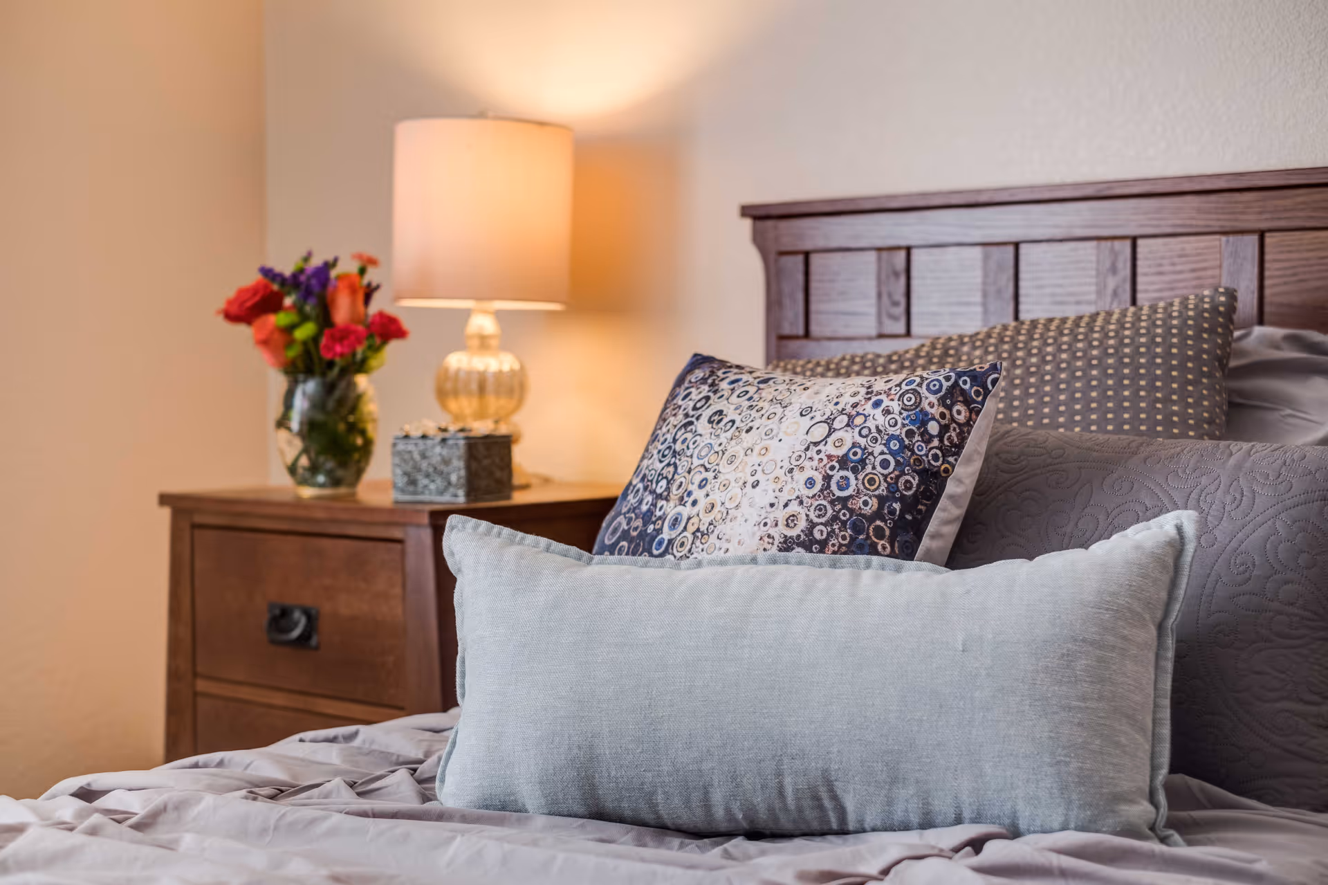 A neatly made bed with decorative pillows and a wooden headboard beside a nightstand holding a lamp and a vase of flowers.