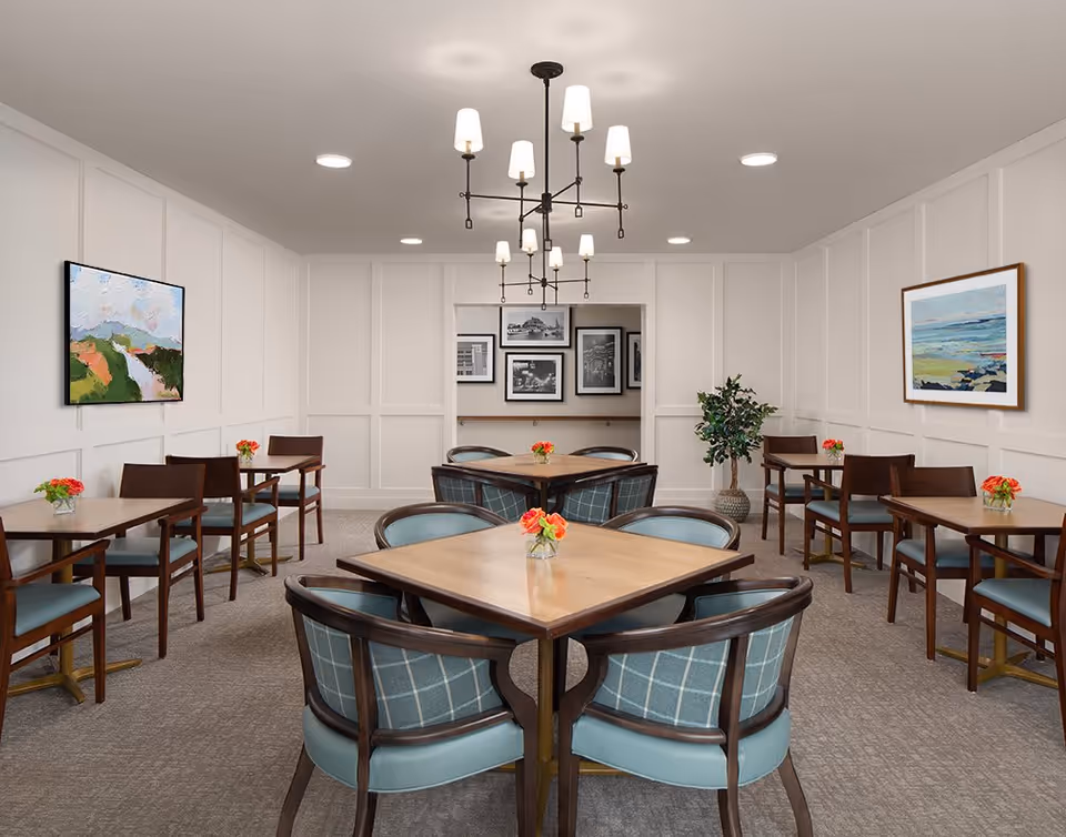 A dining room with multiple wooden tables and chairs arranged neatly. Each table has a small vase with red flowers. The walls are white with paneling and decorated with colorful paintings. A modern chandelier with multiple lights hangs from the ceiling, and there is a potted plant in the corner.