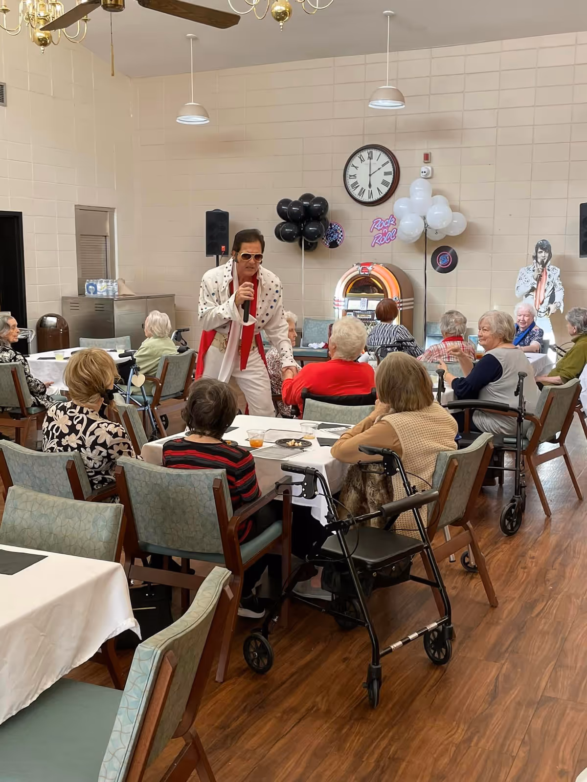 A man dressed as Elvis Presley is performing with a microphone in a senior living facility dining area. Several elderly residents are seated at tables watching the performance. The room has a jukebox, black and white balloons, a clock on the wall, and a cutout of Elvis Presley. Some residents use walkers and wheelchairs.