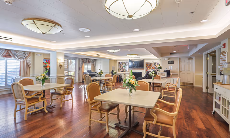 A spacious, well-lit common area with several tables and chairs arranged for seating. Each table has a small floral centerpiece. The room features wooden flooring, large windows with drapes, ceiling lights, and a fireplace with a TV mounted above it. There is a piano in the background and a counter area with a coffee machine on the right side.