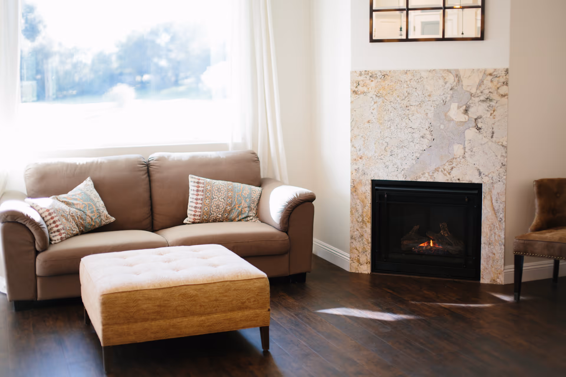 A cozy living room with a beige sofa adorned with two patterned pillows, a matching ottoman in front, a marble fireplace with a small fire burning, and a brown upholstered chair to the right. The room has wooden flooring and a large window with white curtains letting in natural light.