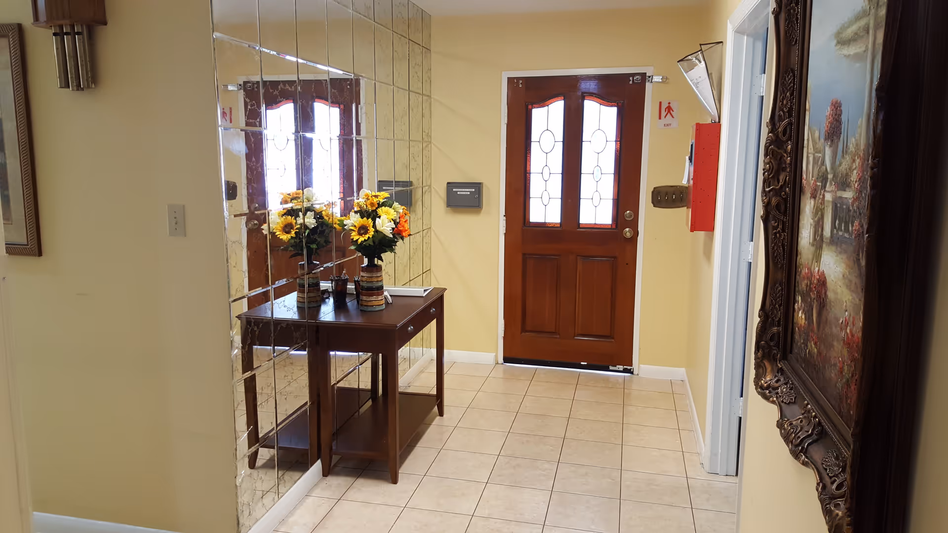 Entryway of a facility with a wooden front door featuring decorative glass panels. A small wooden table with two vases of sunflowers is placed against a mirrored wall on the left. The floor is tiled, and there is a framed painting on the right wall. A fire alarm and exit sign are visible near the door.