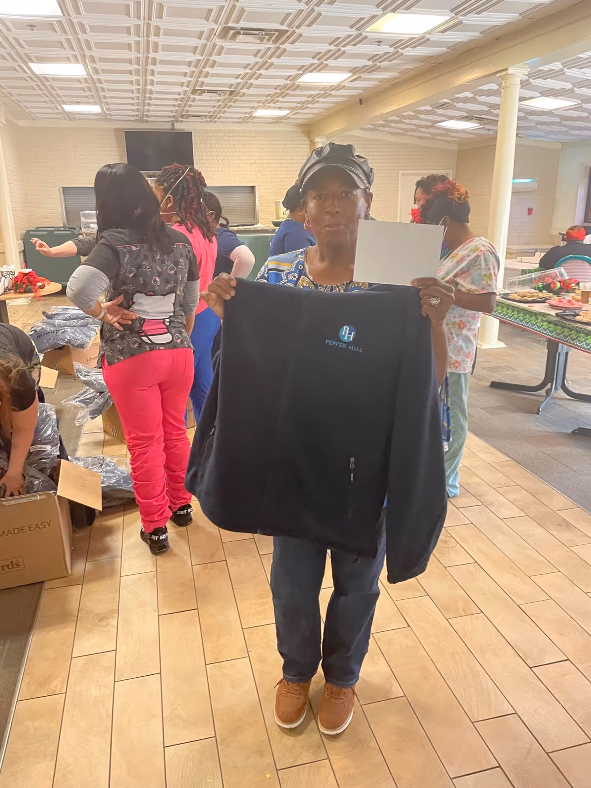 A woman standing indoors holding up a black jacket with the logo 'Pepper Hill' on it and a blank white card. Behind her, several people are engaged in activities such as unpacking boxes and preparing food on tables in a spacious room with tiled flooring and a decorative ceiling.