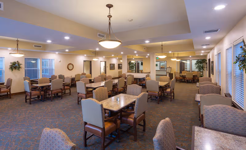 A spacious dining room in a senior living facility with multiple tables and cushioned chairs arranged neatly. The room is well-lit with ceiling lights and pendant lamps, and has large windows with blinds. There are decorative plants and framed pictures on the walls, creating a warm and inviting atmosphere.