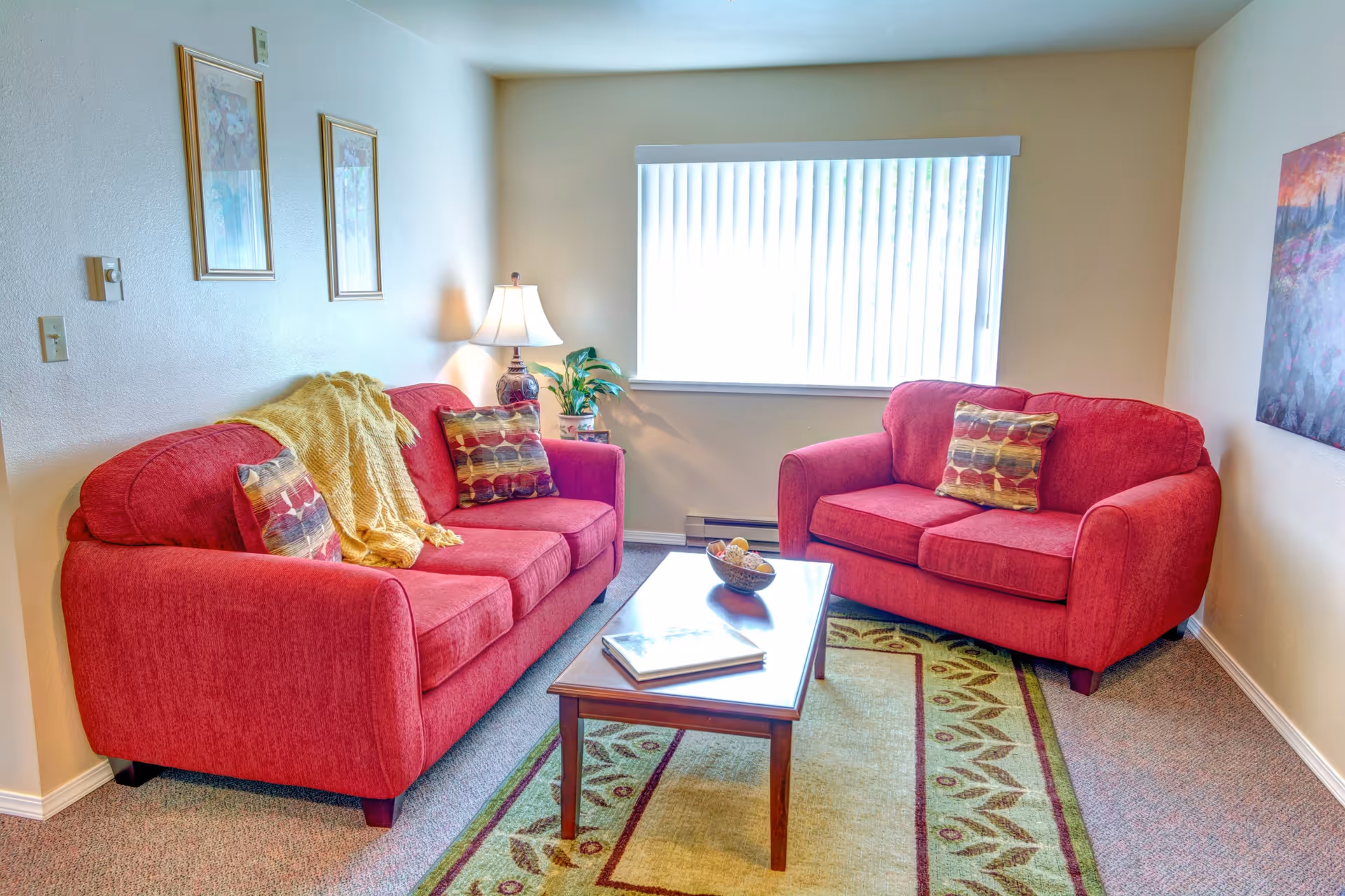 A cozy living room with two red upholstered sofas, each with patterned throw pillows. A wooden coffee table sits between the sofas on a decorative area rug. A table lamp and a potted plant are on a small side table next to one sofa. The room has beige walls with framed artwork and a large window with vertical blinds letting in natural light.