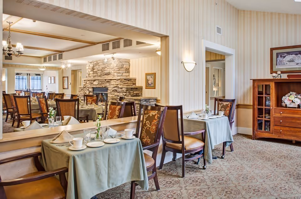 A cozy dining room with tables covered in light green tablecloths, set with white cups, plates, and napkins. The room features wooden chairs with cushioned seats and backs, a large mirror reflecting the space, a stone fireplace, and a wooden cabinet with floral decorations. The walls have light striped wallpaper and the floor is carpeted with a patterned design.