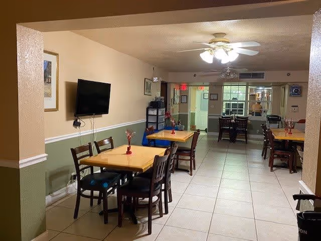 Interior view of a dining area in an assisted living facility with several wooden tables and chairs arranged neatly. A ceiling fan with lights is on, and a flat-screen TV is mounted on the wall. The walls are painted beige and green with white trim, and there are framed pictures hanging. The floor is tiled, and there is a doorway leading to another room in the background.