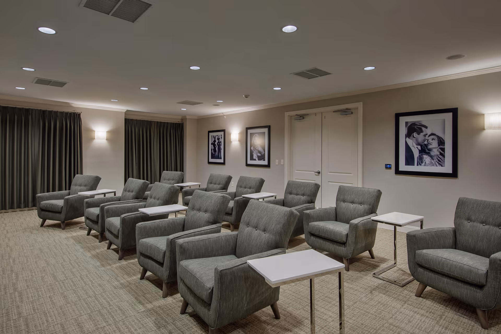Rows of gray upholstered armchairs with small side tables arranged in a quiet lounge with framed black-and-white photos and curtains.
